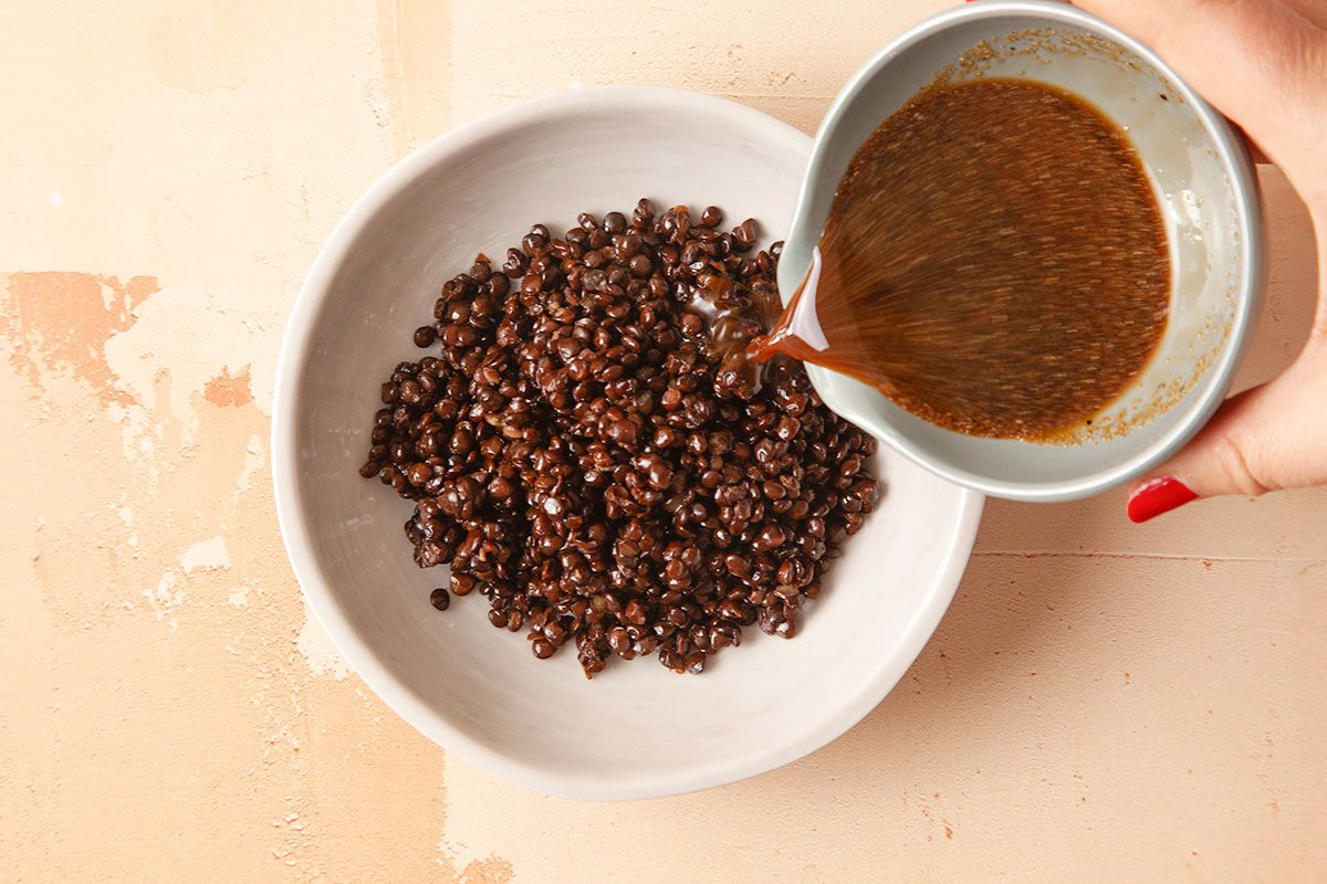 Overhead horizontal image of vinaigrette being poured over cooked black lentils in a mixing bowl.