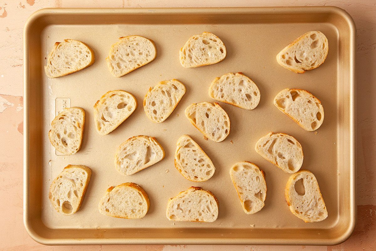 Overhead horizontal image of sliced baguette rounds arranged on a baking sheet ready for toasting.