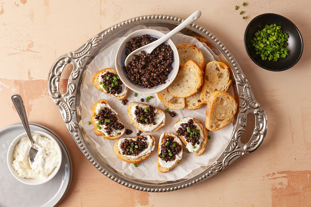 Overhead horizontal image of crostini topped with lentil mixture and garnished with fresh chives on a serving tray.