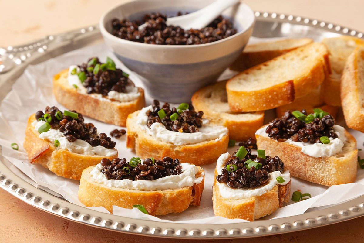 Eye-level horizontal close-up of crostini topped with lentil caviar and fresh chives on a serving tray.