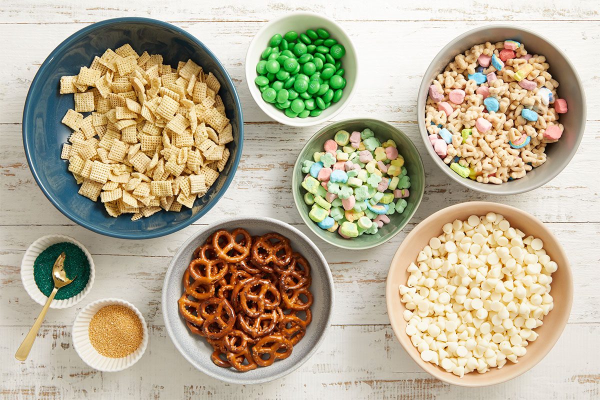 Overhead step-by-step image showing the ingredients for Leprechaun Bait snack mix arranged in bowls on a light wood surface. The ingredients include Rice Chex cereal, Lucky Charms cereal, miniature pretzels, green M&M’s, Lucky Charms marshmallows, white chocolate chips, and green and gold sanding sugar.