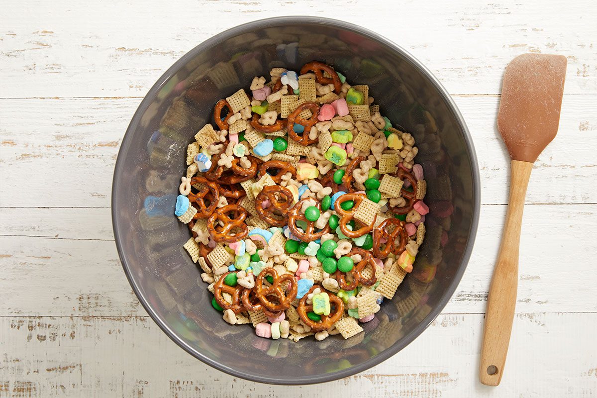 Overhead step-by-step image showing cereal, pretzels, marshmallows, and green chocolate candies combined in a large mixing bowl as the Leprechaun Bait snack mix begins to come together.