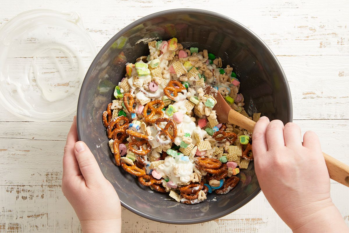 Overhead step-by-step image showing hands stirring the Leprechaun Bait mixture in a large mixing bowl with a wooden spoon to combine the cereal, pretzels, candies, and marshmallows.