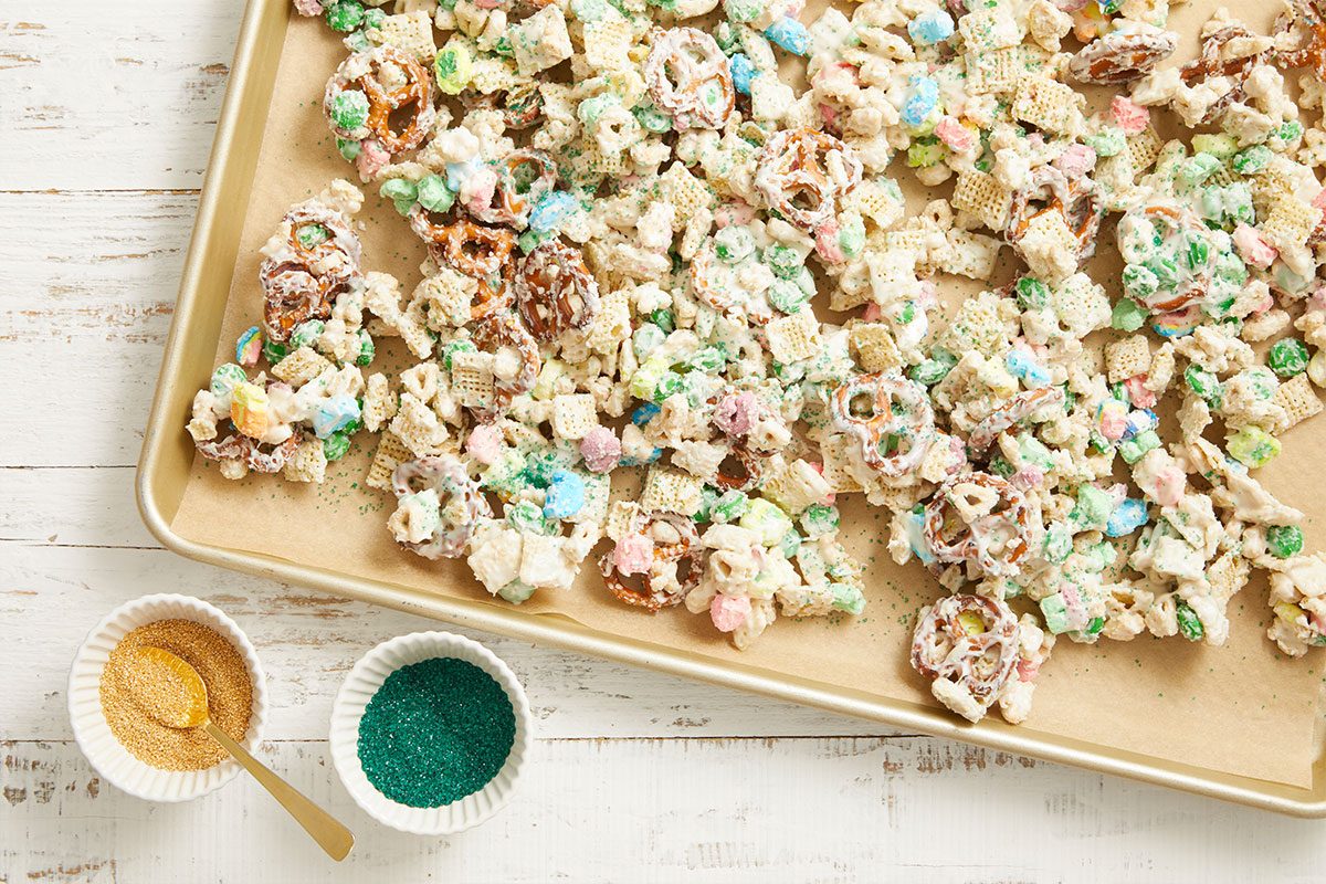 Overhead step-by-step image showing the Leprechaun Bait snack mixture spread across a baking sheet and sprinkled with green and gold sanding sugar for a festive St. Patrick’s Day treat.