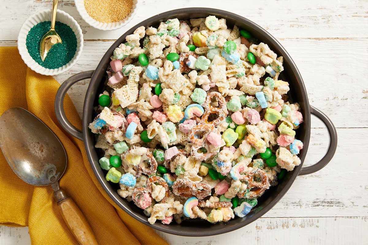 Push-and-pull overhead image showing a serving bowl filled with Leprechaun Bait snack mix featuring cereal, pretzels, marshmallows, and colorful candies.