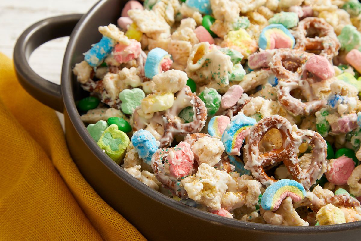 Close-up push-and-pull image showing a detailed view of Leprechaun Bait snack mix with cereal pieces, pretzels, marshmallows, candies, and white chocolate coating.