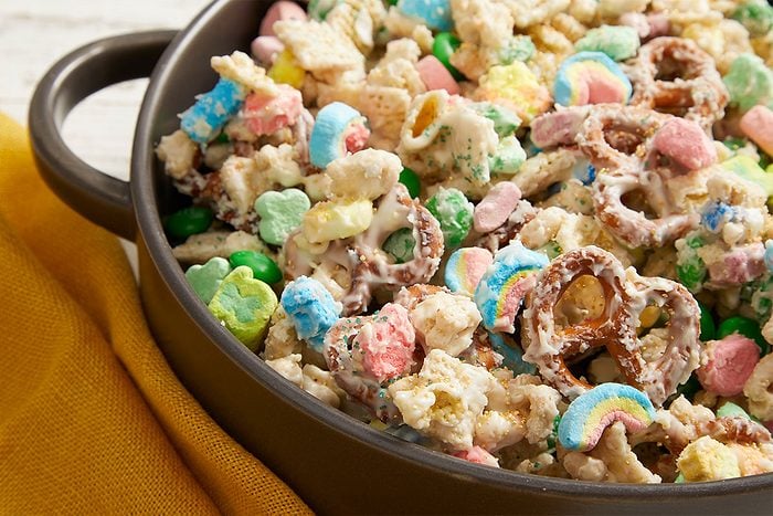 Close-up push-and-pull image showing a detailed view of Leprechaun Bait snack mix with cereal pieces, pretzels, marshmallows, candies, and white chocolate coating.