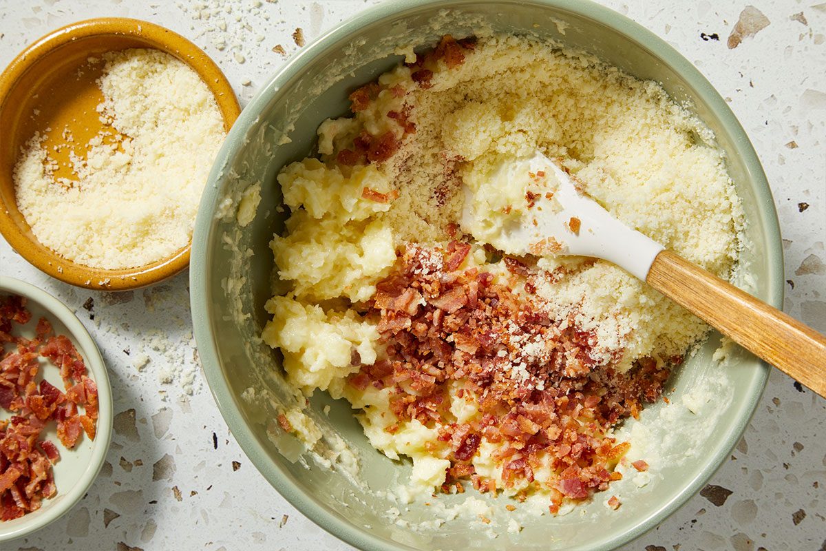 Overhead shot of mashed potatoes combined with bacon bits, cheese, and seasonings in a bowl, stirred with a spatula.