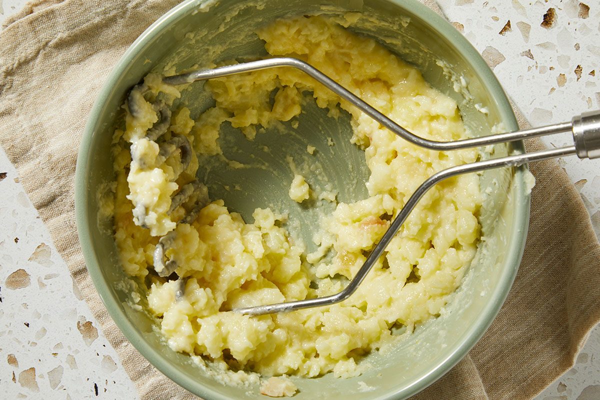 Overhead shot of scooped potato flesh being mashed in a bowl, with butter and seasonings mixed in using a fork.
