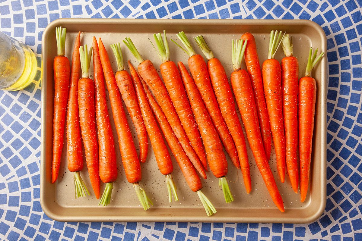 Overhead view of fresh whole carrots with green tops arranged in a baking sheet before roasting. The carrots are lined up neatly on a tray against a blue patterned background, showing the raw ingredients used for the miso-roasted carrot recipe.