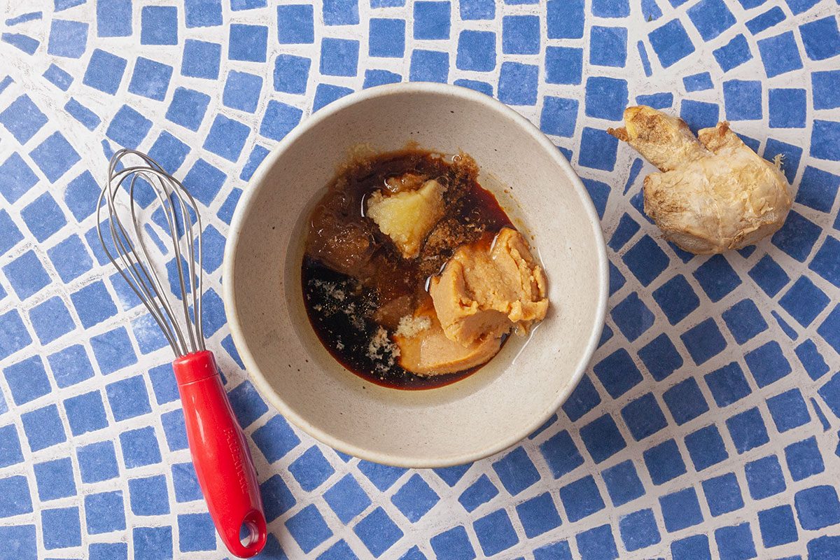 Overhead shot of a bowl containing miso paste, soy sauce, brown sugar, sesame oil, minced ginger and garlic being whisked together to create a savory glaze for roasted carrots. A whisk rests beside the bowl with fresh ginger nearby, illustrating the sauce preparation for the dish.