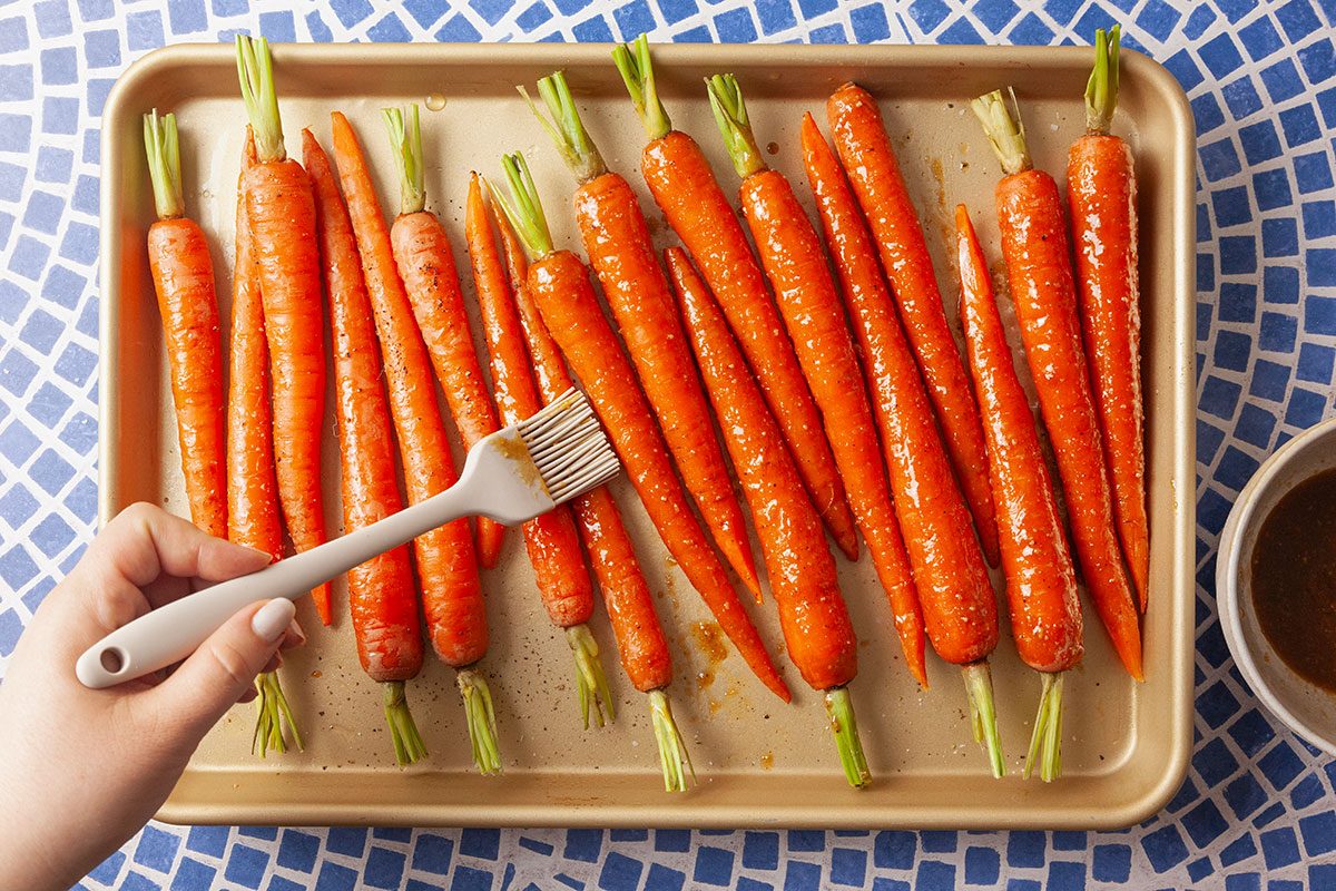 Overhead view of fresh carrots arranged on a baking sheet as a hand reaches in with a fork to begin coating or preparing the vegetables for roasting. The carrots are lined neatly on the tray, illustrating the preparation stage before cooking.