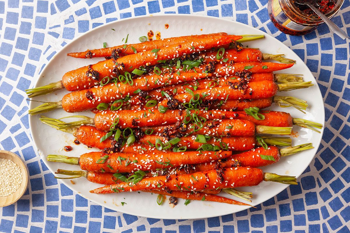 Overhead view of roasted carrots arranged on a white platter and coated in a rich miso glaze. The carrots are garnished with sesame seeds, chopped cilantro, sliced green onions and chili crisp for a finished presentation of the dish.