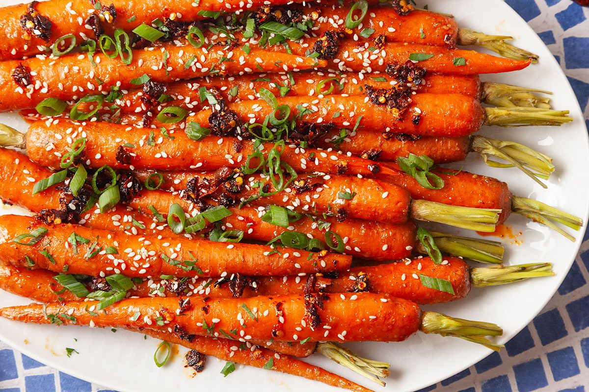 Close-up overhead view of miso-roasted carrots on a serving platter highlighting the glossy glaze and garnish of sesame seeds, chopped herbs and green onions. The vibrant orange carrots are arranged tightly together for a colorful presentation.