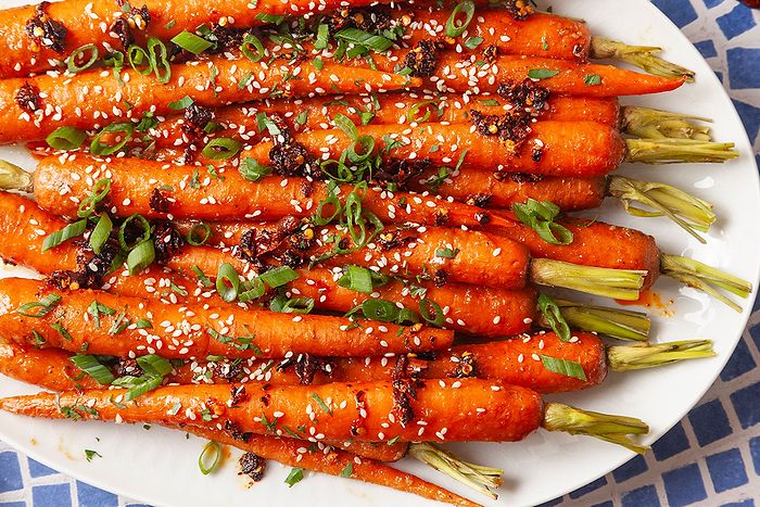 Close-up overhead view of miso-roasted carrots on a serving platter highlighting the glossy glaze and garnish of sesame seeds, chopped herbs and green onions. The vibrant orange carrots are arranged tightly together for a colorful presentation.