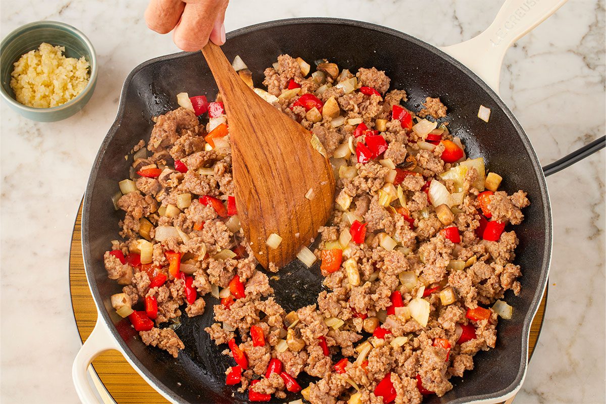 Overhead shot of a hand stirring ground meat, chopped onions, and red bell peppers in a cast-iron skillet with a wooden spatula, with a small bowl of minced garlic nearby on a marble countertop;