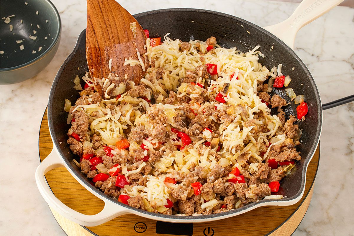 Close-up shot of a skillet on a stovetop containing cooked ground meat, chopped red bell peppers, onions, and shredded cheese, being stirred with a wooden spatula;
