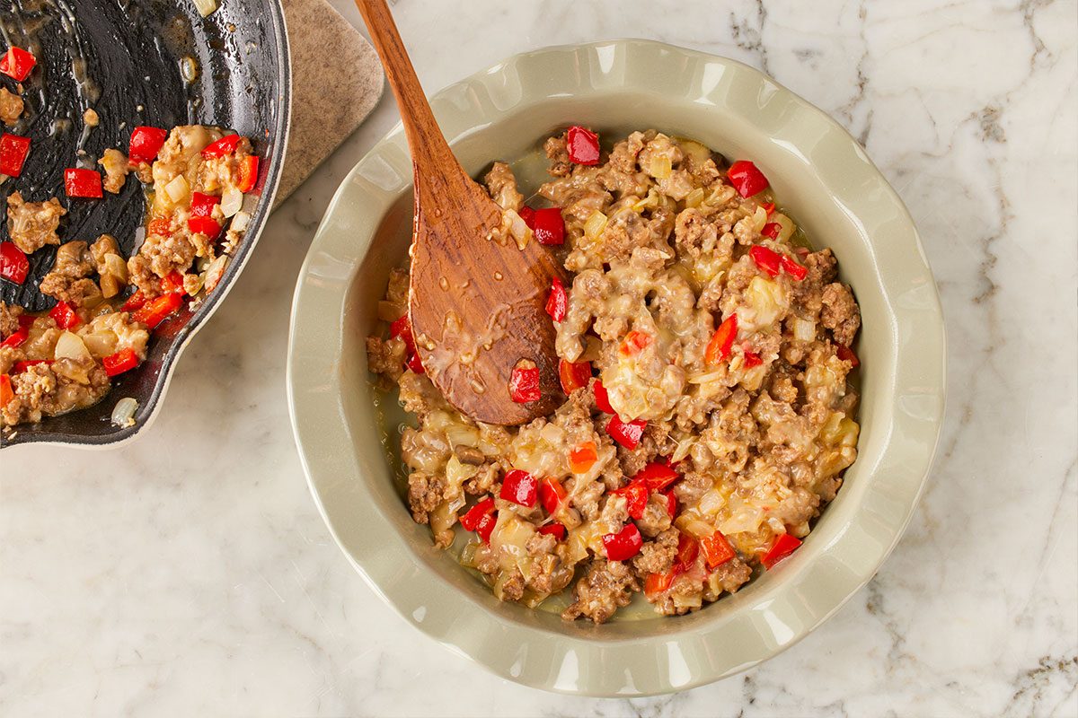 Overhead shot of a ceramic dish filled with a mixture of ground meat, diced red bell peppers, and onions, with a wooden spoon resting inside; a skillet with more of the mixture sits nearby on a marble countertop;