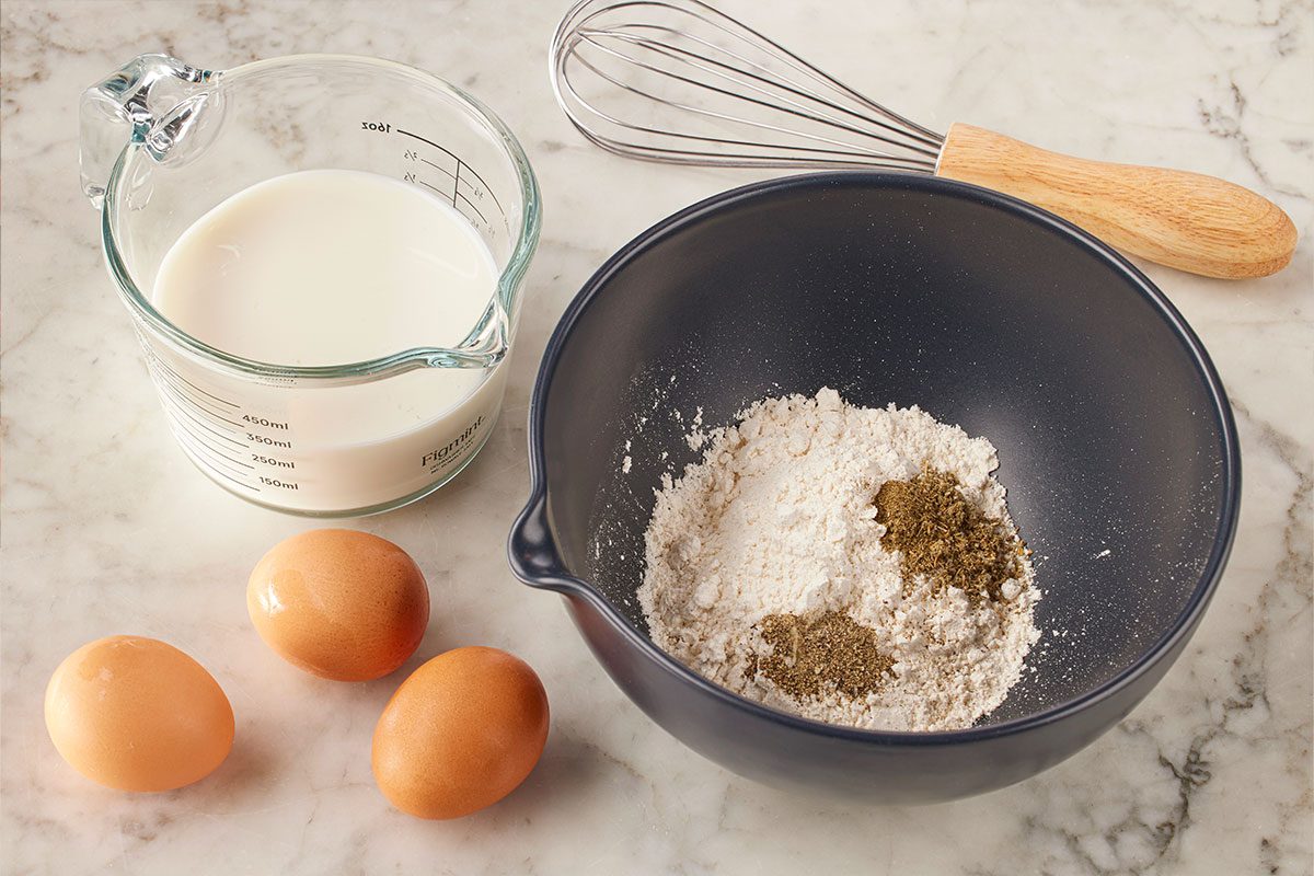 Overhead shot of a whisk, a measuring cup of milk, three eggs, and a mixing bowl containing flour and spices, arranged on a marble countertop;