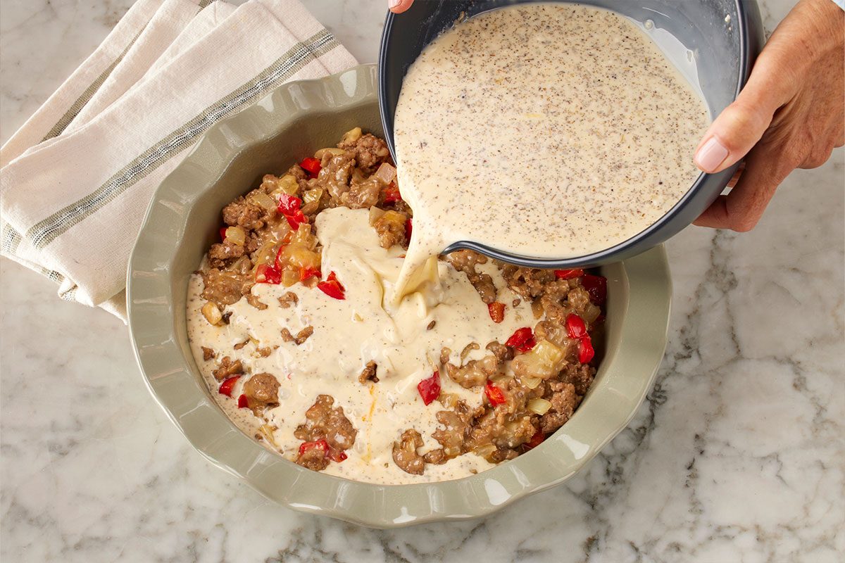 Overhead shot of a hand pouring a creamy egg mixture from a bowl over a casserole dish filled with sausage, potatoes, and red peppers, on a marble countertop with a folded napkin nearby;