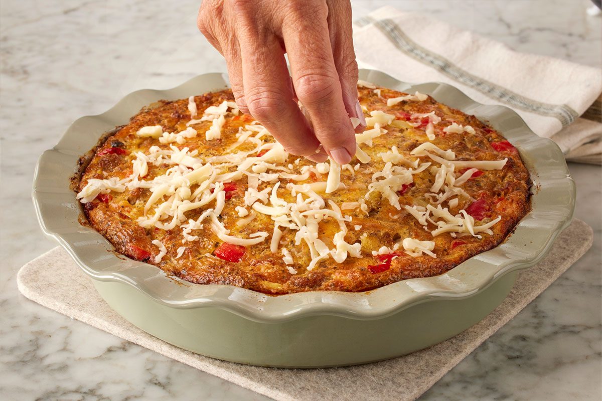 Table-view shot of a hand sprinkling shredded cheese over a baked quiche with red and green vegetables, in a round ceramic dish on a marble countertop;