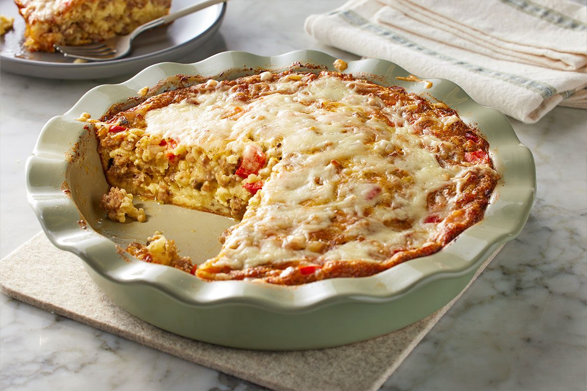 Table view shot of Monterey Sausage Pie with melted cheese in a round dish, a slice removed to reveal eggs, sausage, and vegetables, with a serving plate and fork placed behind;