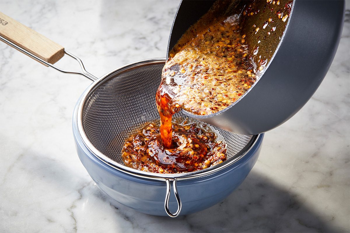 Overhead horizontal image of hot honey sauce being strained into a bowl, showing chili flakes and spices.