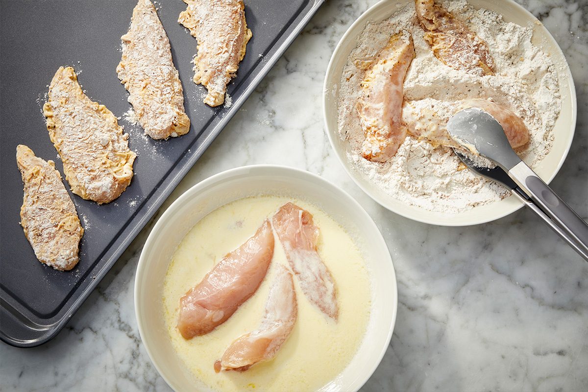 Overhead horizontal image of chicken tenders being dredged in flour and dipped in egg and buttermilk mixture.