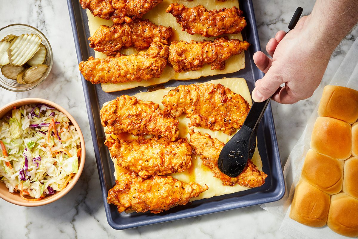 Overhead horizontal image of fried chicken tenders on a sheet pan being brushed with hot honey glaze next to slider buns and coleslaw.