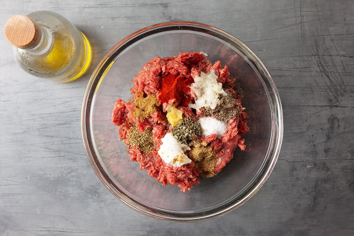Overhead view of a glass mixing bowl filled with ground beef topped with grated onion, Greek yogurt, olive oil, and a blend of spices including oregano, cumin, coriander, paprika, parsley, salt and pepper. Ingredients are arranged on a gray surface, ready to be mixed for homemade gyros.