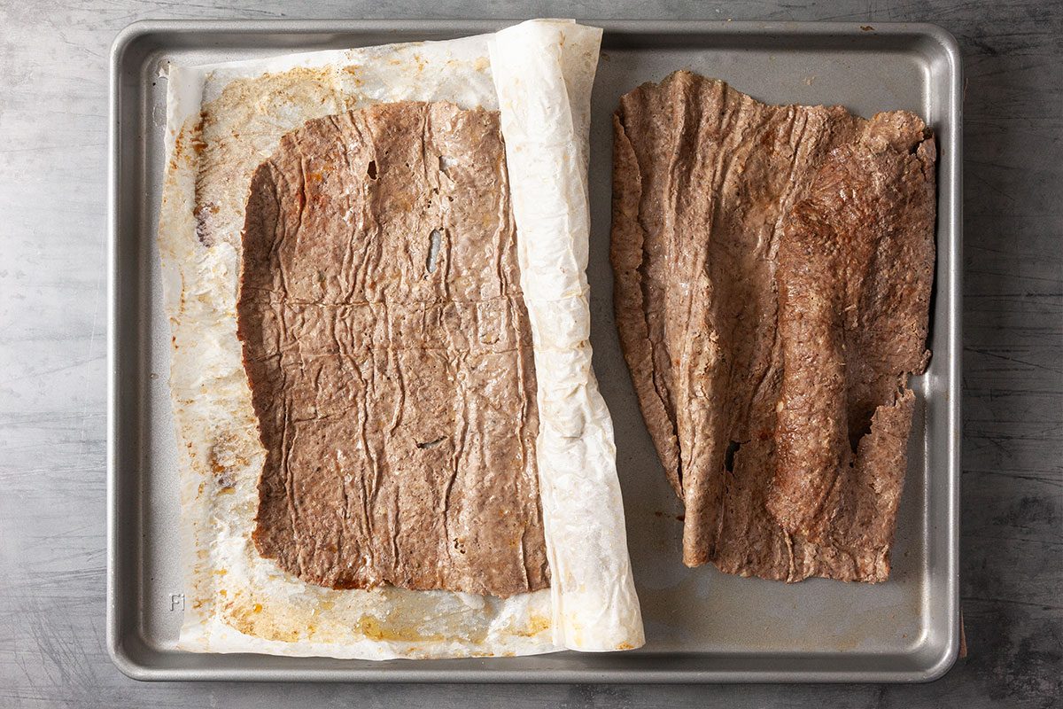 Overhead view of baked gyro meat unwrapped from parchment paper on a sheet pan. The cooked beef slab is browned and firm, ready to be sliced.