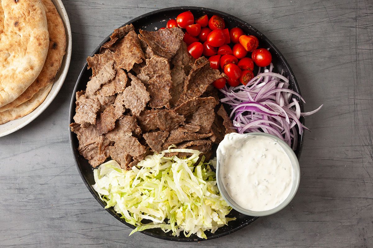 Overhead image of a platter featuring sliced gyro meat served with shredded lettuce, cherry tomatoes, sliced red onion and a bowl of tzatziki sauce alongside warm flatbreads.
