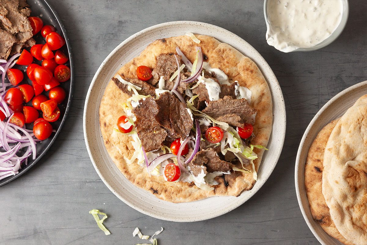 Overhead image of a fully assembled gyro on a plate topped with sliced beef, shredded lettuce, tomatoes and red onion, served with tzatziki sauce on the side.