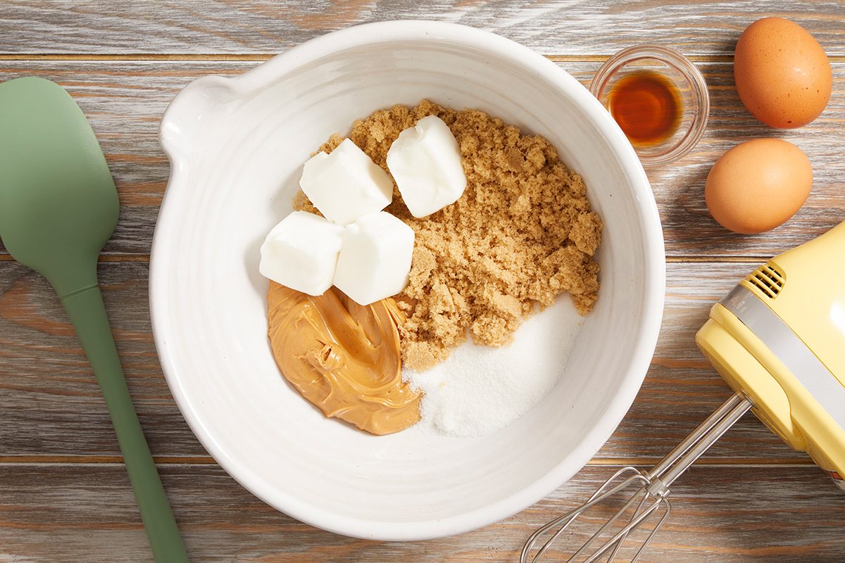 A mixing bowl with brown sugar, white sugar, peanut butter, and cubed butter, next to a spatula, vanilla extract, two eggs, and a hand mixer on a wooden surface.
