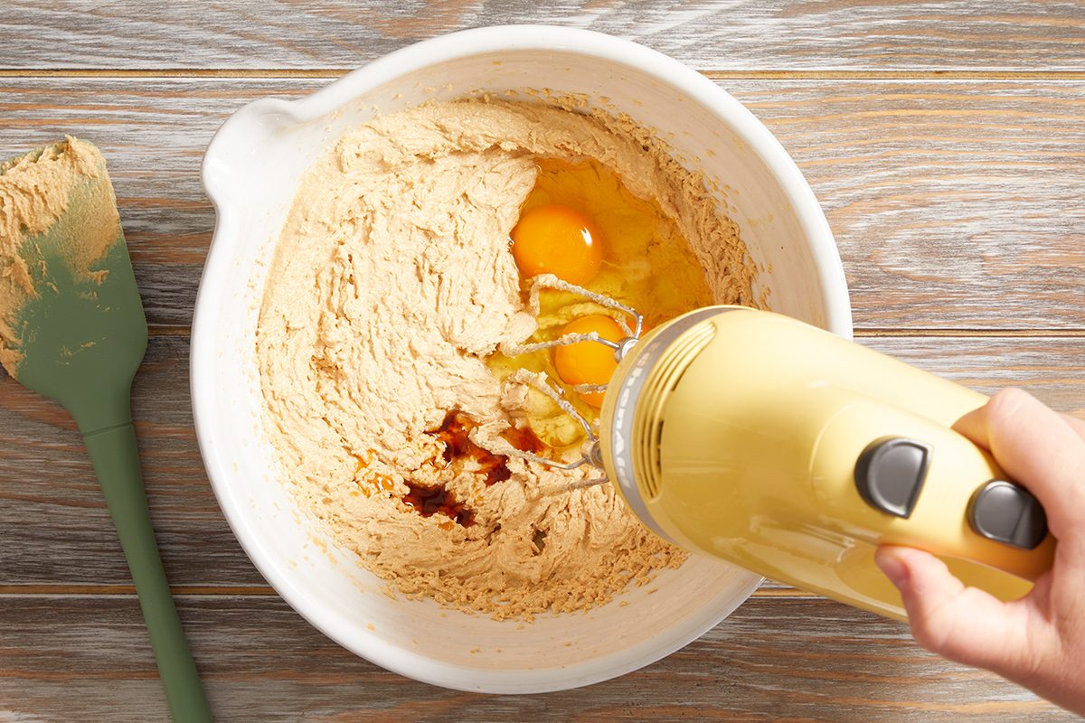 A person uses a yellow electric hand mixer to blend eggs and vanilla into a bowl of cookie dough. A green spatula with dough on it rests beside the bowl on a wooden surface.