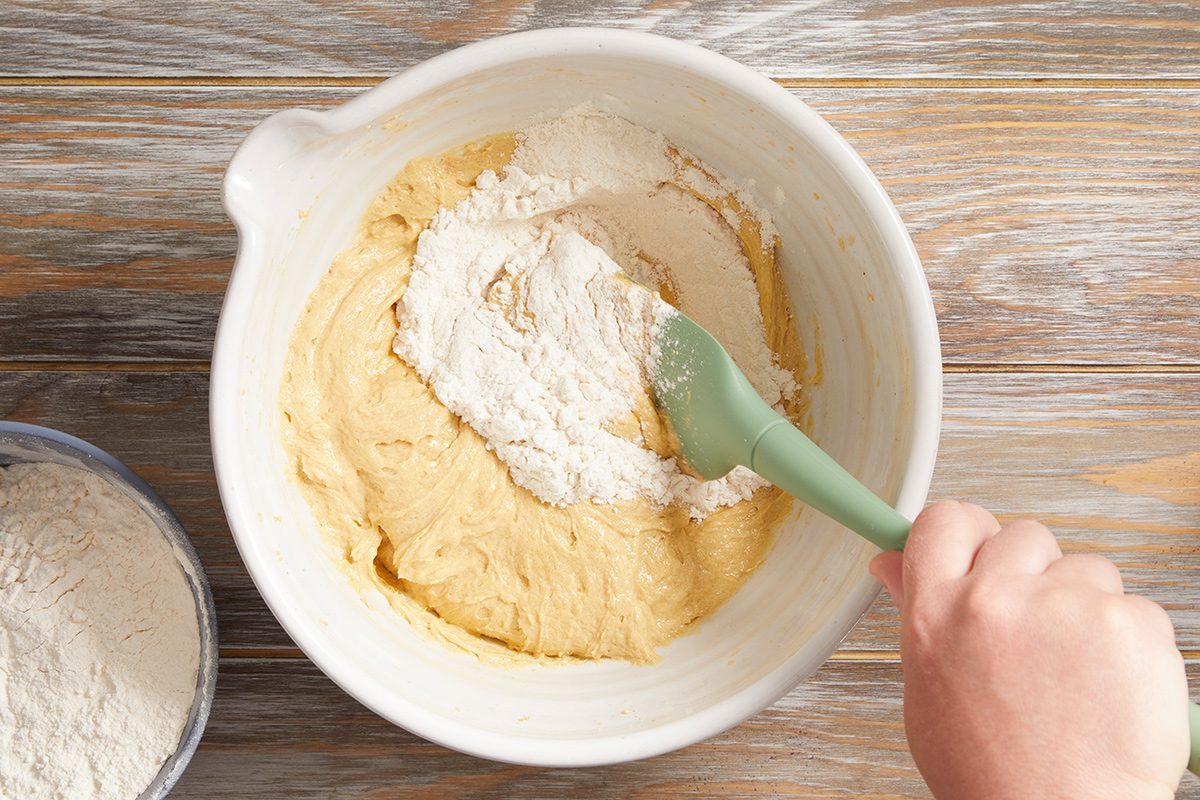 A hand uses a green spatula to mix flour into yellow cake batter in a white mixing bowl on a wooden surface. A small bowl of flour sits nearby.