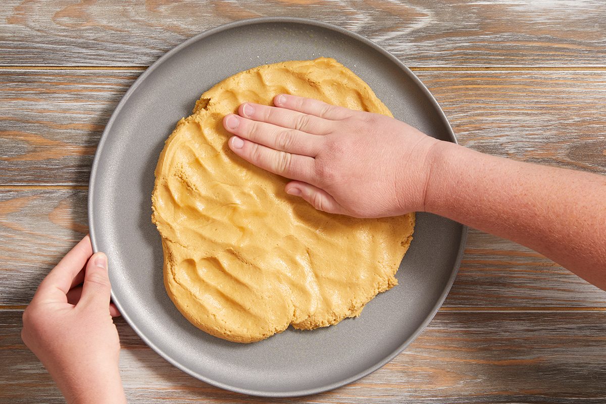 A person presses cookie dough flat onto a round, gray plate with one hand while holding the plate steady with the other hand on a wooden surface.