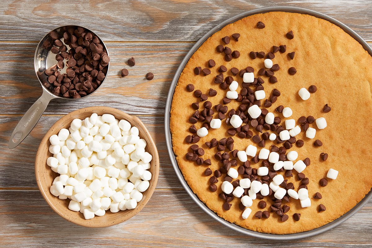 A large cookie on a round baking tray is topped with mini marshmallows and chocolate chips; beside it are a bowl of mini marshmallows and a measuring cup of chocolate chips on a wooden surface.