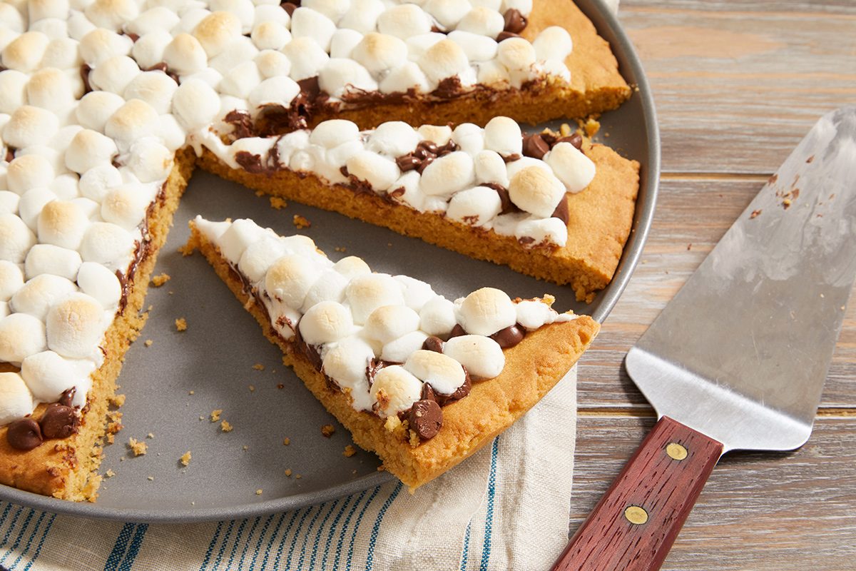 A large round cookie topped with melted chocolate and mini marshmallows is sliced, with one piece removed and a pie server resting nearby on a wooden surface.
