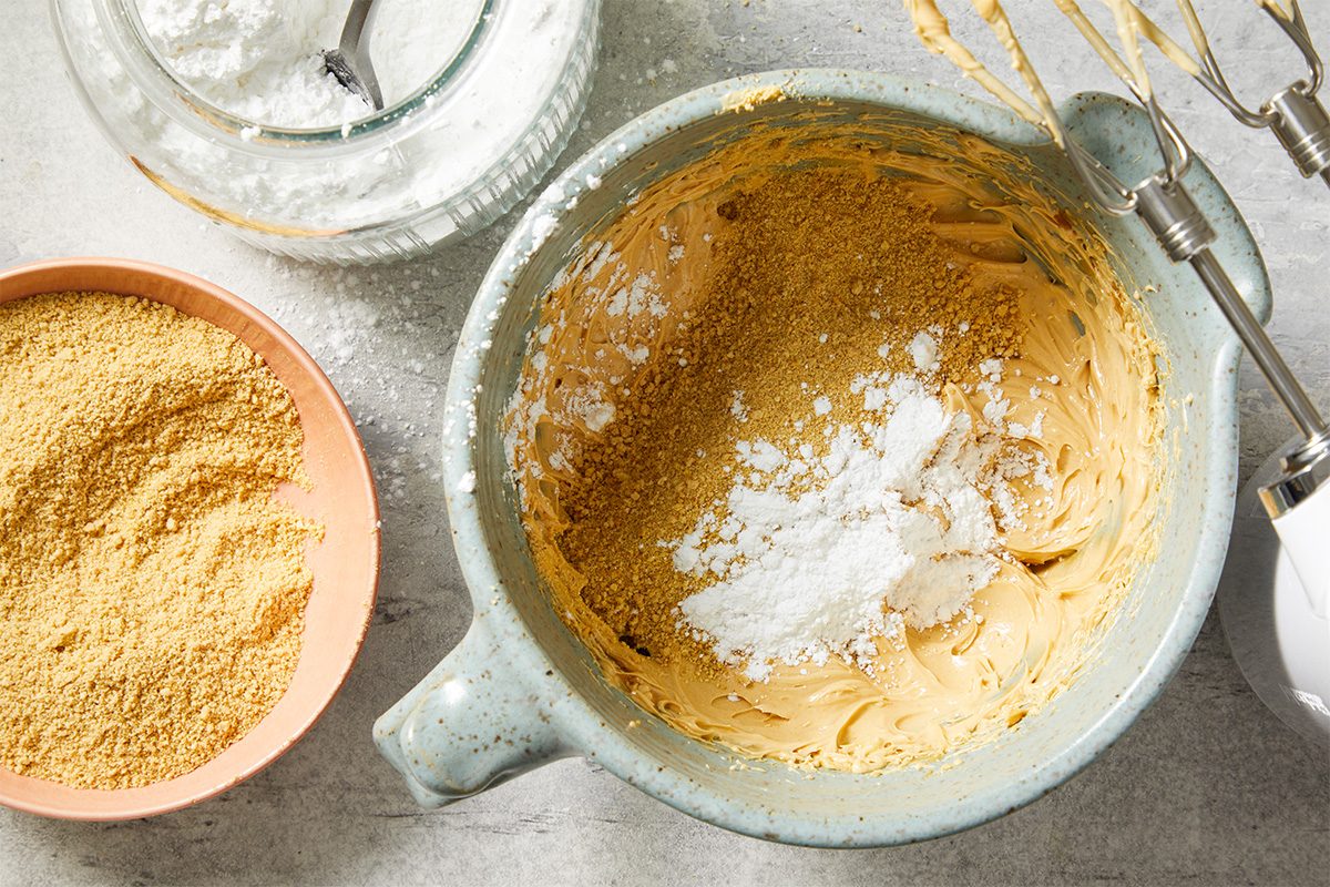 Overhead view of a mixing bowl filled with creamy peanut butter and softened butter as confectioners’ sugar is added. Graham cracker crumbs sit nearby, illustrating the preparation stage for Peanut Butter Robin Eggs candy centers.