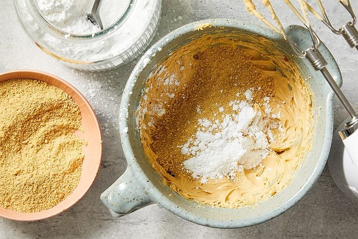 Overhead view of a mixing bowl filled with creamy peanut butter and softened butter as confectioners’ sugar is added. Graham cracker crumbs sit nearby, illustrating the preparation stage for Peanut Butter Robin Eggs candy centers.