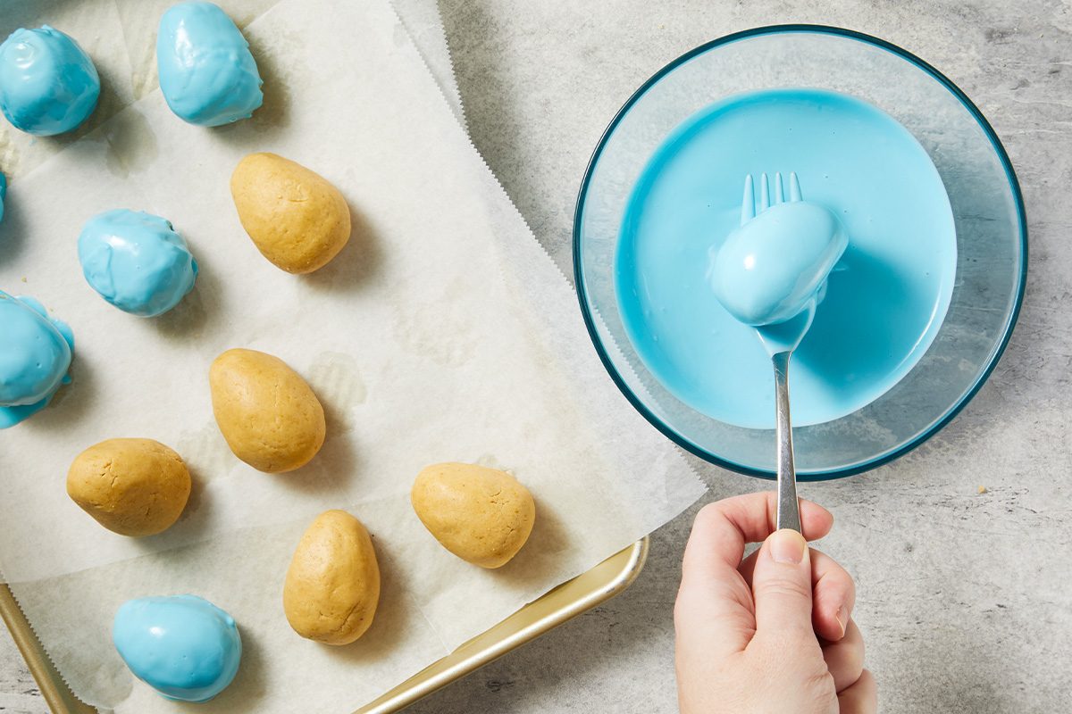 Overhead action shot of peanut butter egg centers being dipped into melted light blue candy coating. A fork lifts a coated egg from the bowl, capturing the candy-dipping process.