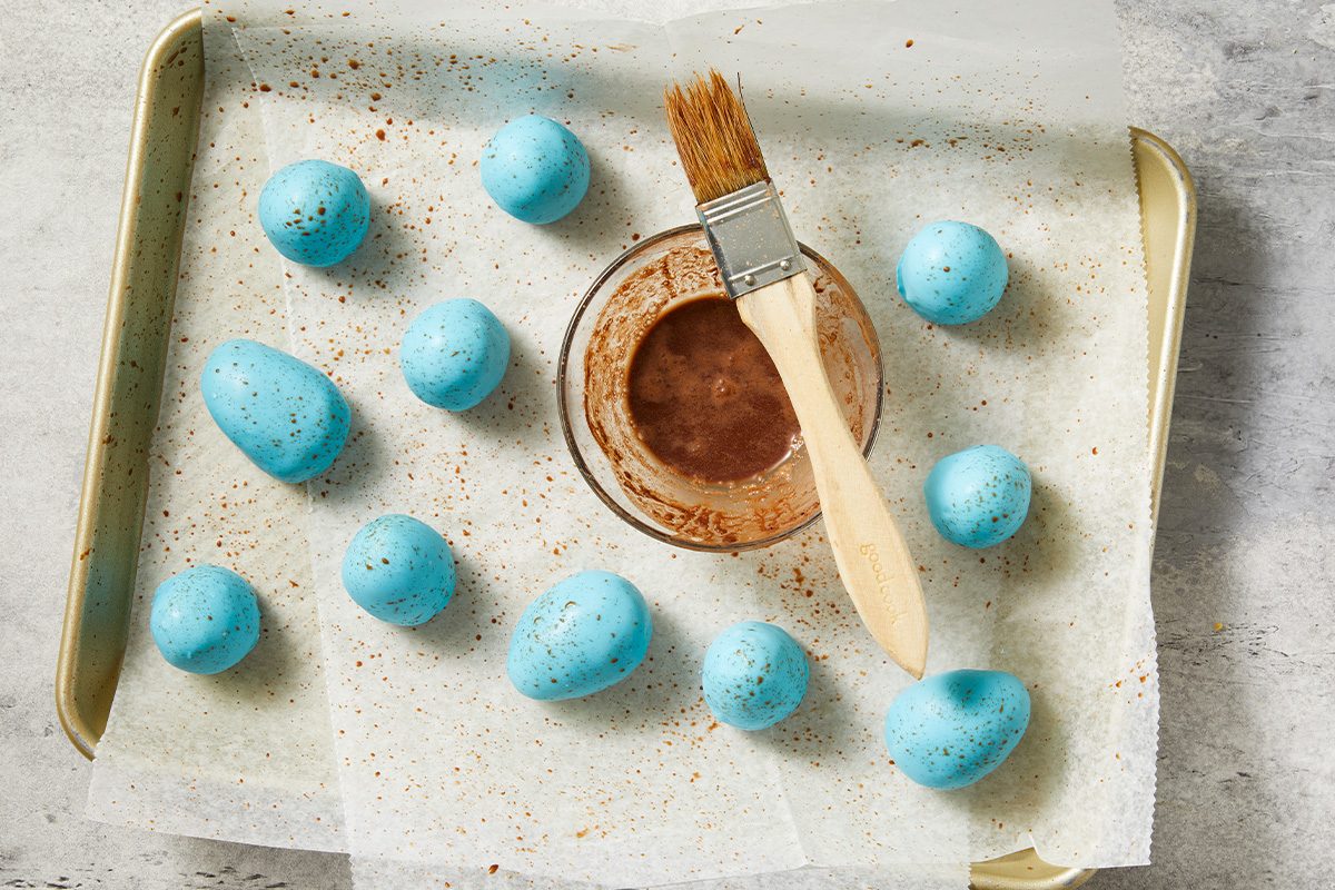 Overhead view of blue candy-coated peanut butter eggs arranged on a lined baking sheet while cocoa speckles are brushed on for a robin egg effect. A pastry brush and cocoa mixture are visible, demonstrating the finishing detail.