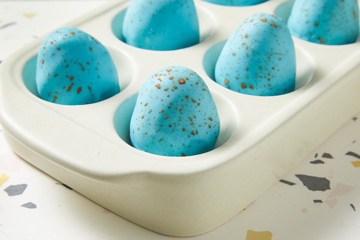 Horizontal close-up of Peanut Butter Robin Eggs nestled in a serving dish, highlighting the smooth blue coating and cocoa speckled finish. The image emphasizes texture and presentation.