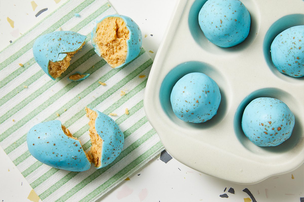 Horizontal overhead image of Peanut Butter Robin Eggs with one candy cut open to reveal the creamy peanut butter interior. The contrast between the blue candy shell and golden filling showcases texture and flavor.