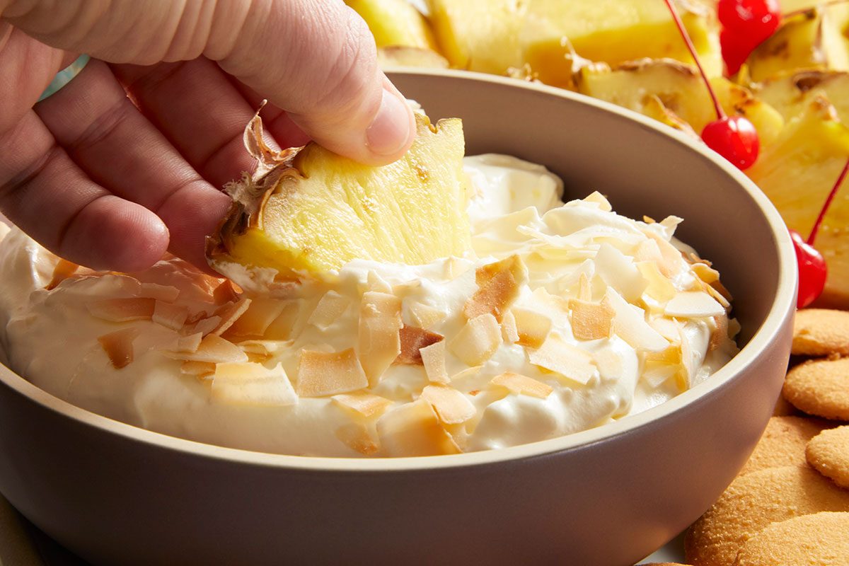 Close-up shot of a hand dipping a pineapple wedge into the creamy Pina Colada Dip