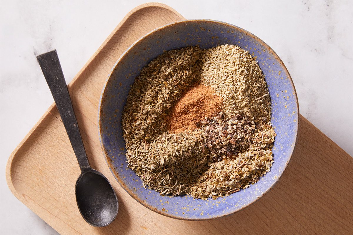 Overhead shot of a small blue bowl filled with ground poultry seasoning mix, with a spoon resting inside and small bits of herbs visible.