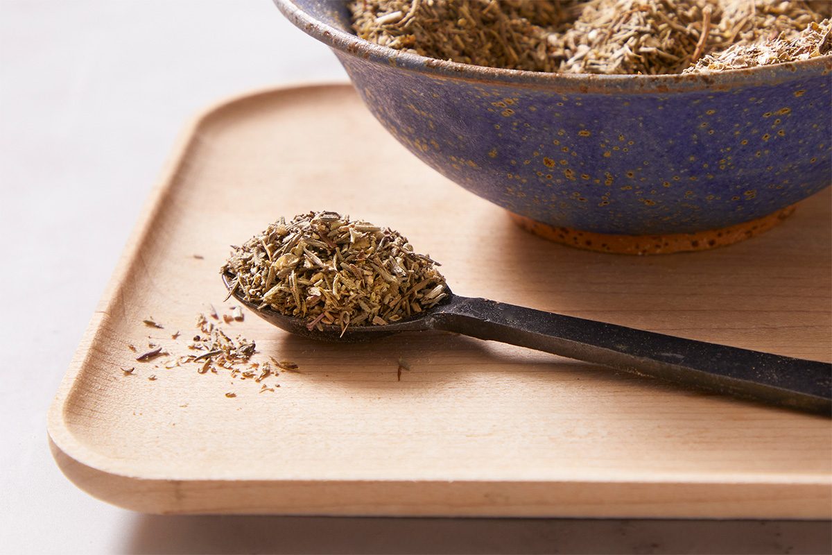 Close-up of poultry seasoning mix in a deep blue bowl on a tray, with a spoonful of seasoning resting beside it on the light wooden surface.
