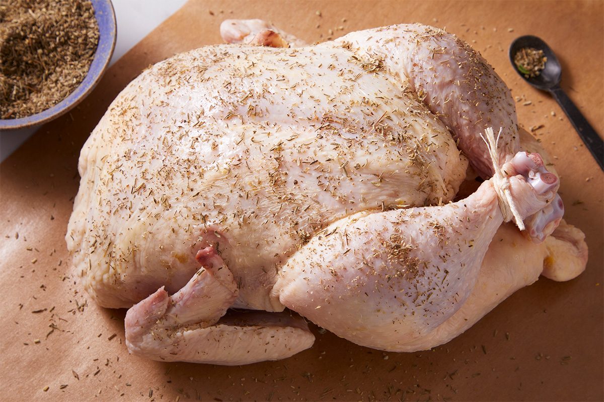 Close-up shot of a whole raw chicken on a wooden board, rubbed with Poultry Seasoning, with a bowl of seasoning mix in the background.