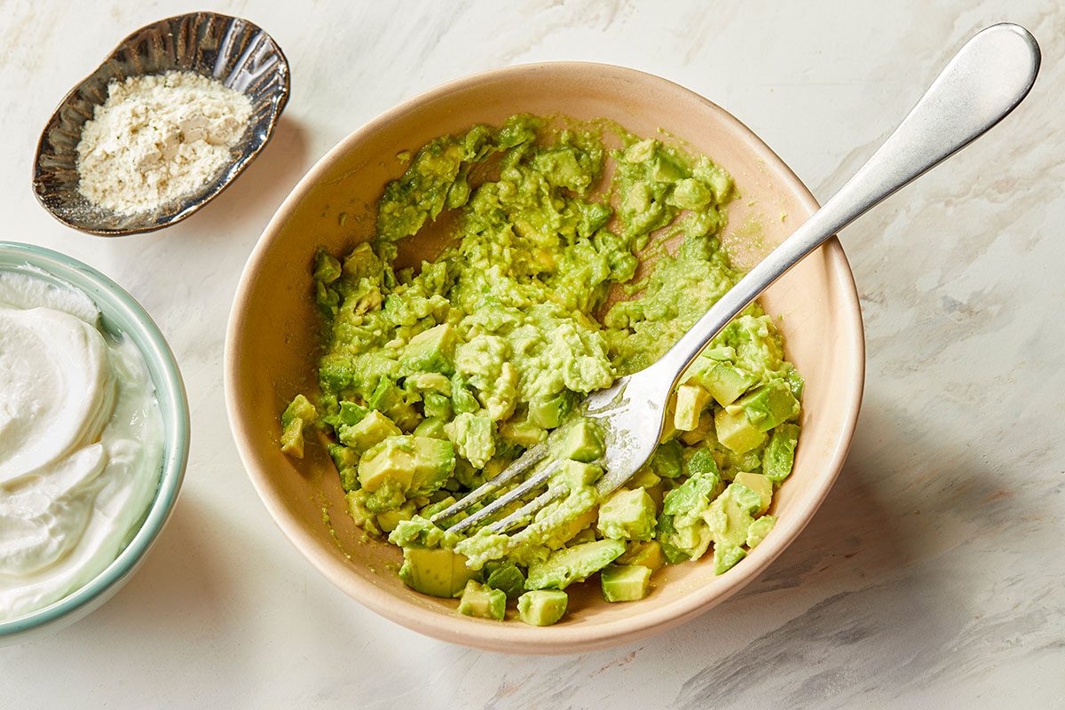 Overhead image of diced avocado being mashed in a bowl with lime juice to begin preparing Ranch Avocado Yogurt Dip.